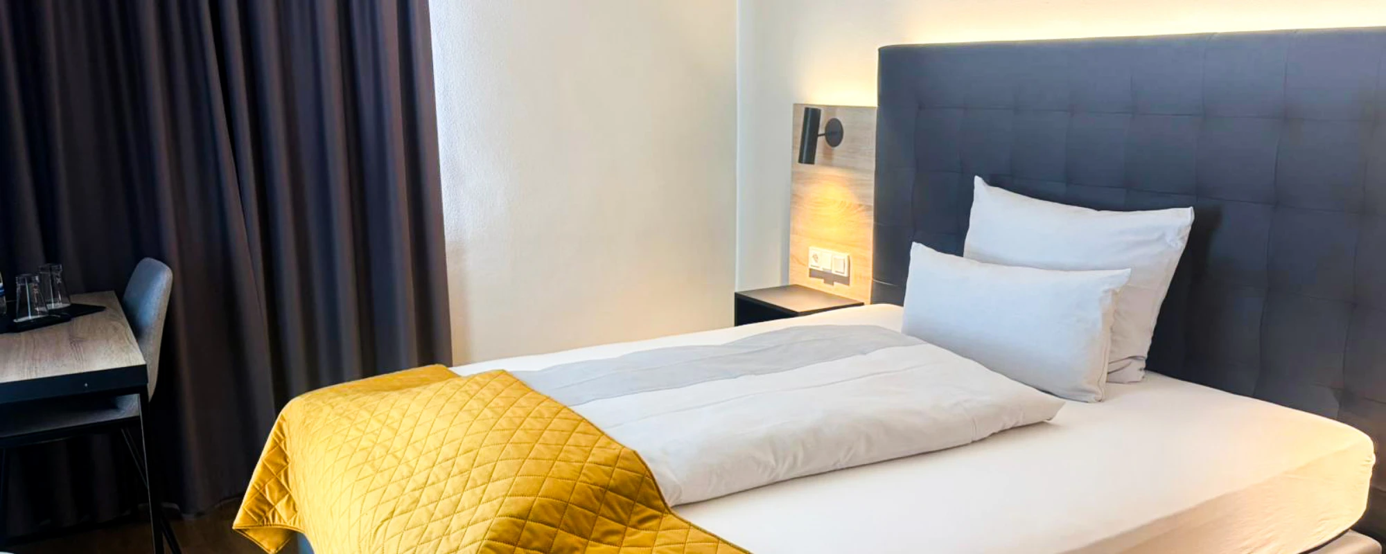 Einzelzimmer mit großem Bett, dunkelblauem Kopfteil, gelber Steppdecke am Fußende, Schreibtisch mit Stuhl und dunklen Vorhängen