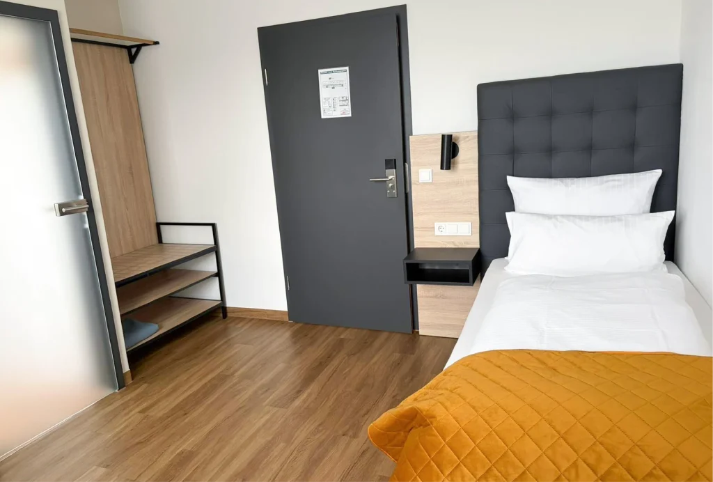 Einzelzimmer mit Bett, grauer Tür, Holzboden, Wandregal und Nachttisch mit Steckdosen.