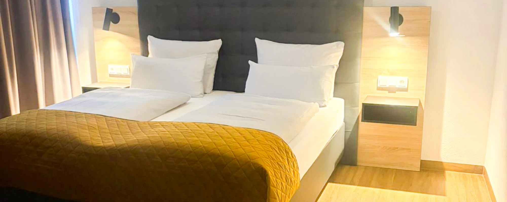 Doppelbett mit gepolstertem Kopfteil, zwei weißen Kissen, gelber Steppdecke und zwei hölzernen Nachttischen mit Wandlampen in einem hellen Hotelzimmer