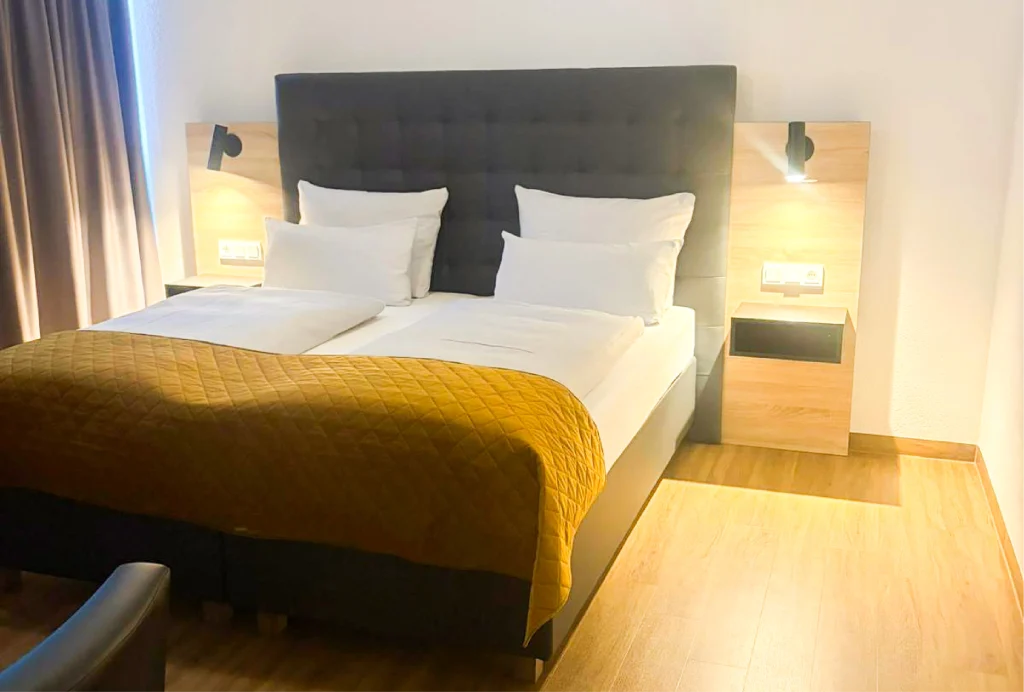 Doppelbett mit gepolstertem Kopfteil, zwei weißen Kissen, gelber Steppdecke und zwei hölzernen Nachttischen mit Wandlampen in einem hellen Hotelzimmer