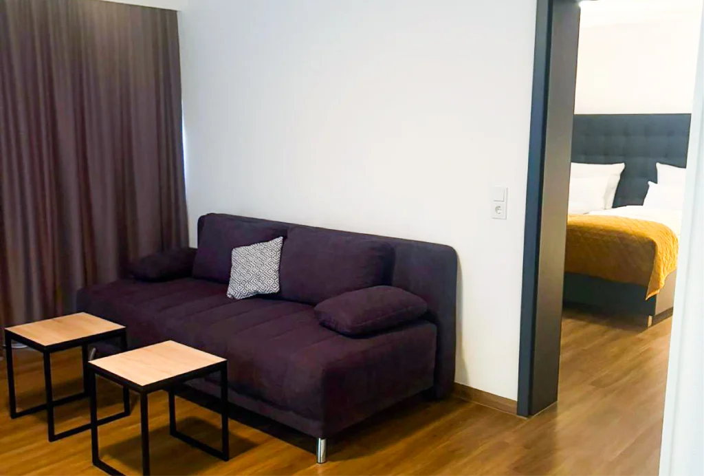 Dunkelviolette Couch mit zwei Kissen vor weißen Wänden und zwei kleinen quadratischen Tischen mit Holzplatten auf Holzfußboden, daneben Türöffnung zu Schlafzimmer mit Bett und gelber Decke