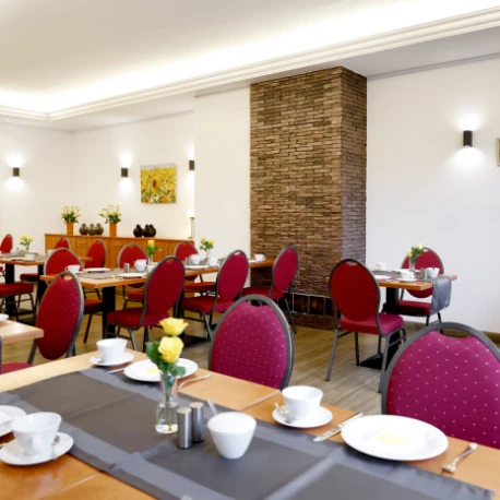 Frühstücksraum mit mehreren gedeckten Tischen, roten gepolsterten Stühlen, einer Steinwand als Raumteiler und weißen Wänden mit Wandlampen und einem Blumenbild