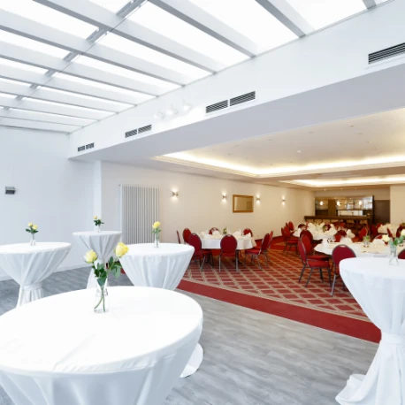 Großer heller Saal mit Tischen in weißer Tischdecke und roten Stühlen auf gemustertem Teppichboden unter einer Glasdecke
