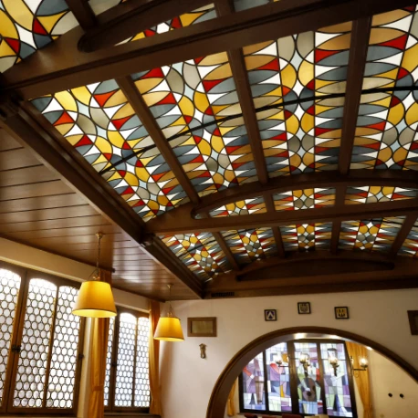 Decke mit bunten Glasfenstern in geometrischem Muster und Holzbalken in einem Raum mit runden Fenstern und gelben Hängelampen
