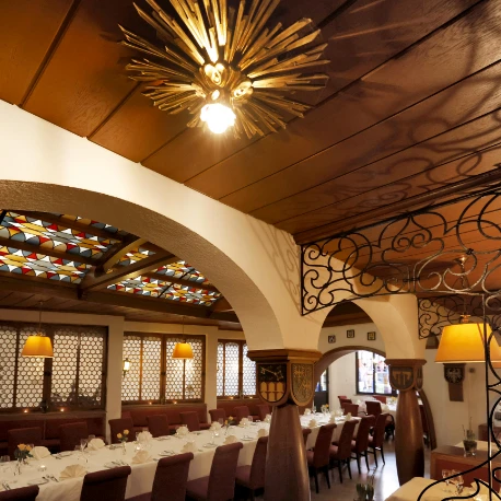 Langer festlich gedeckter Tisch in einem historischen Rittersaal mit Holzdecke, verzierten Säulen und einem großen Kronleuchter mit Sonnenstrahlen-Design