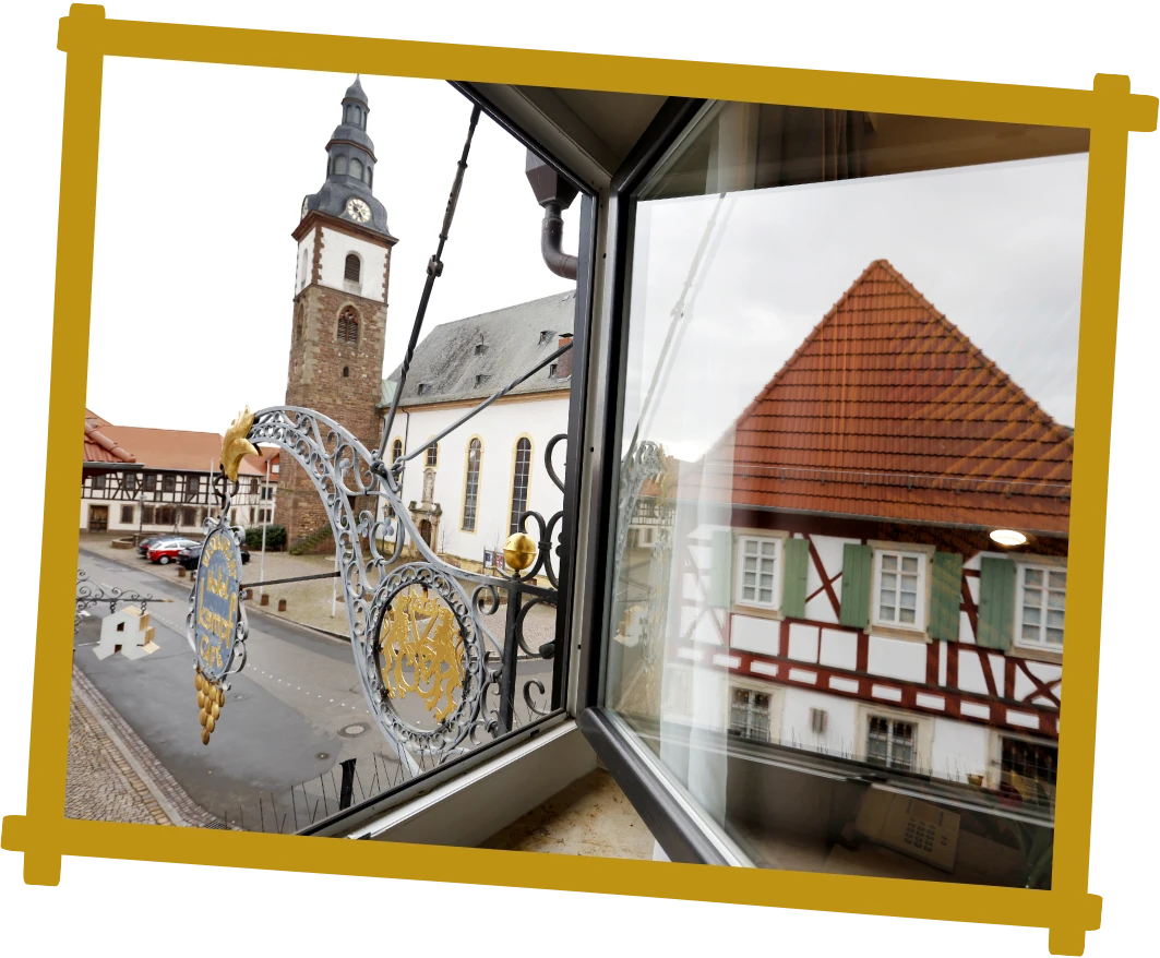 Blick aus einem geöffneten Fenster auf eine historische Kirche mit Turm, ein Fachwerkhaus und ein kunstvoll verziertes, hängendes Schild an einer Straße