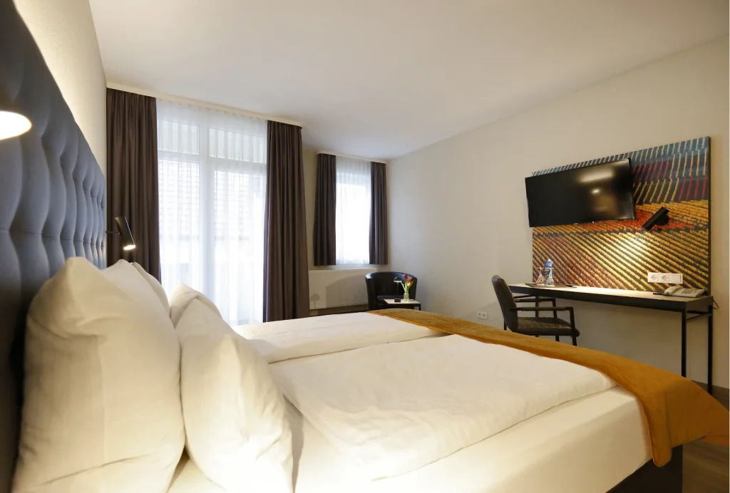 Hotelzimmer mit zwei Einzelbetten, großem Fenster mit Vorhängen, Schreibtisch mit Stuhl und an der Wand montiertem Fernseher
