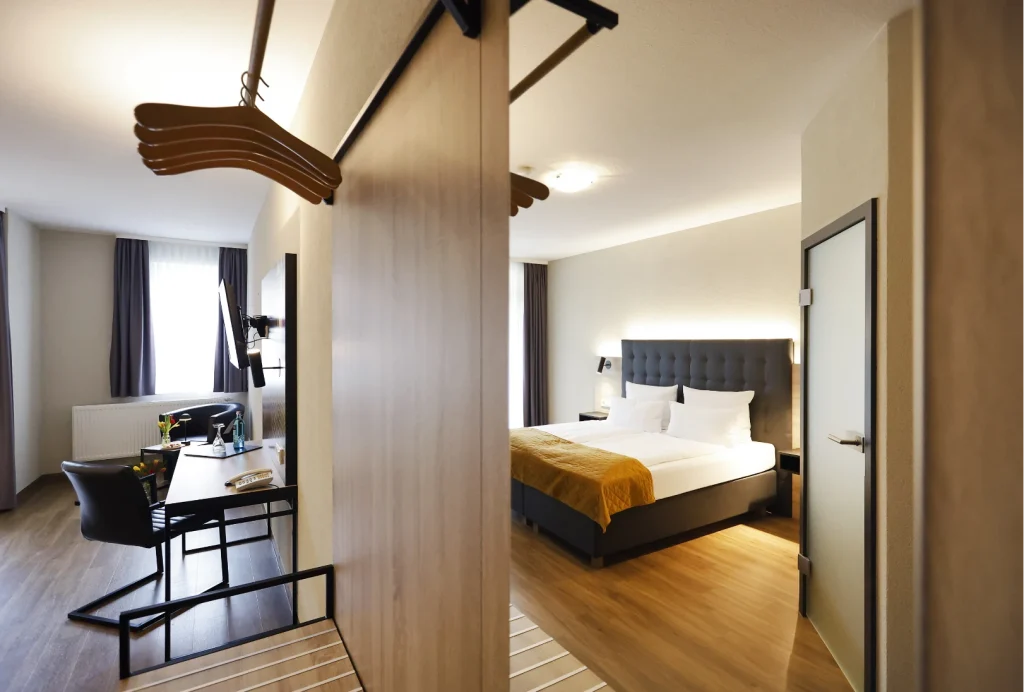 Hotelzimmer mit Doppelbett, Schreibtisch, Stuhl und offenem Kleiderschrank mit Holzkleiderbügeln