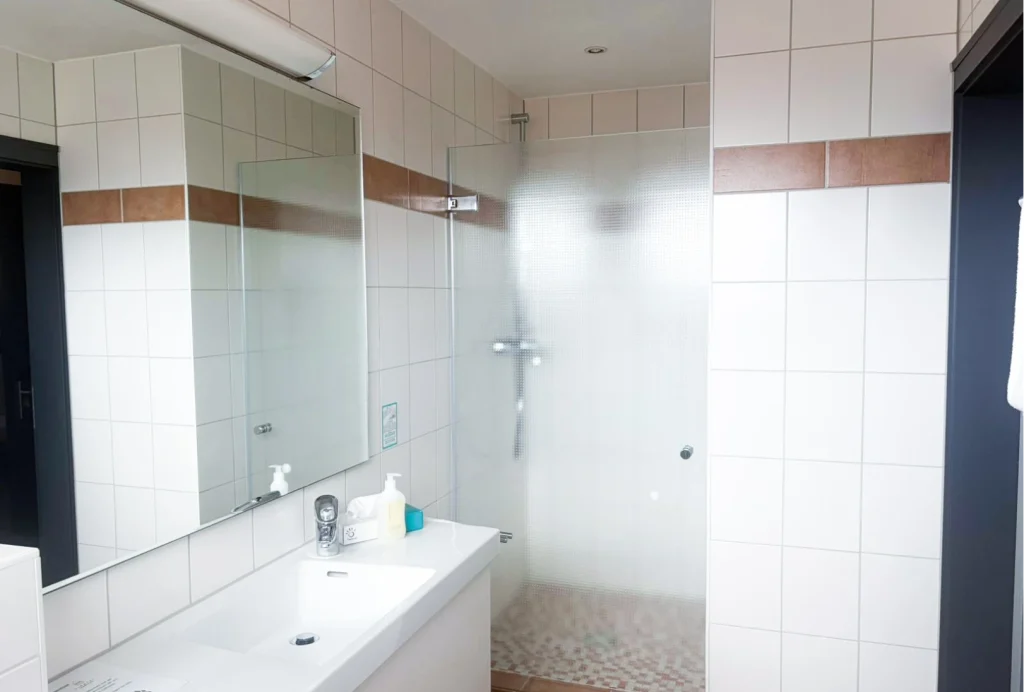 Badezimmer mit Waschbecken, großem Spiegel, Seifenspender und Duschkabine mit milchglasartiger Tür