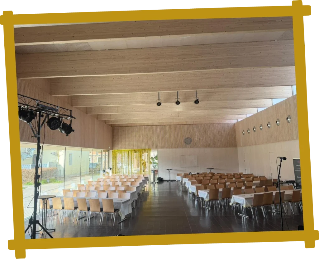 Festhalle mit Reihen von Tischen und Stühlen, Holzbalkendecke und Bühnenbeleuchtung